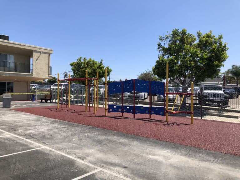 Red rubber-surfaced recreation area with a shaded play structure beside apartment buildings