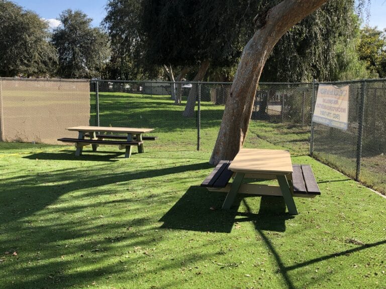 Fenced dog park with synthetic turf, a shade tree and picnic tables