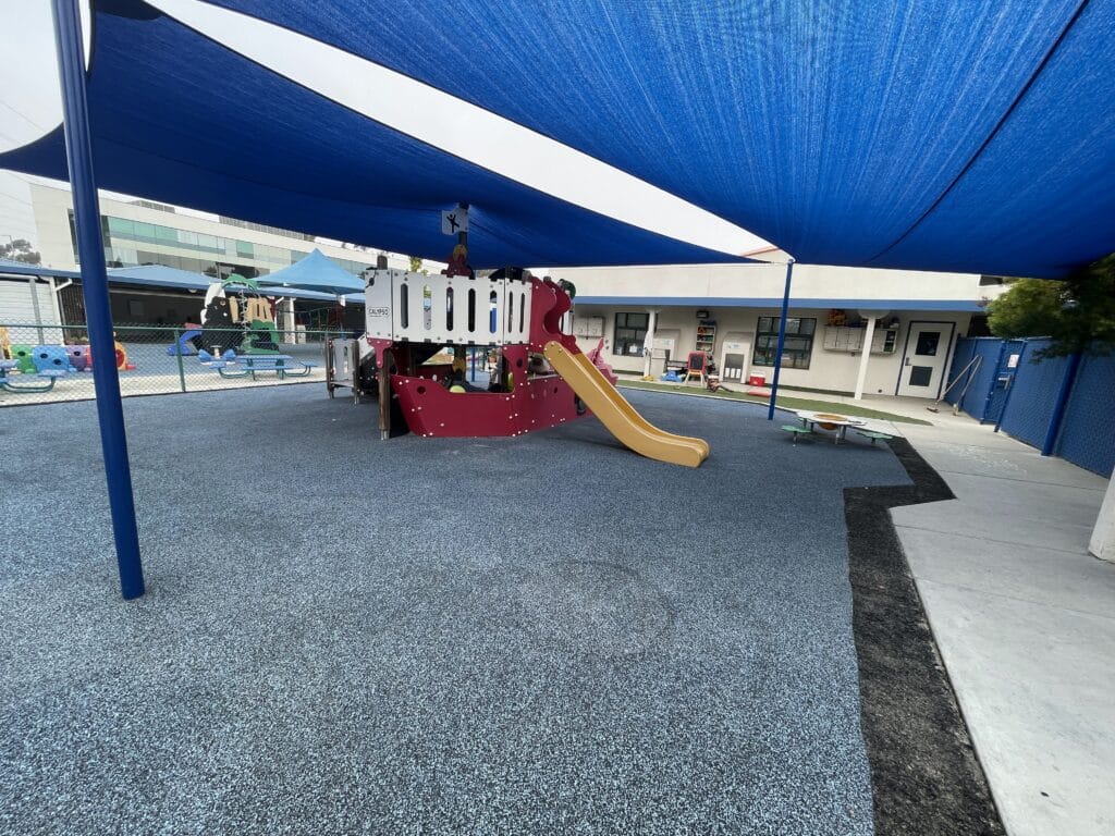 Gray poured-in-place rubber under a shade sail with a red slide play structure