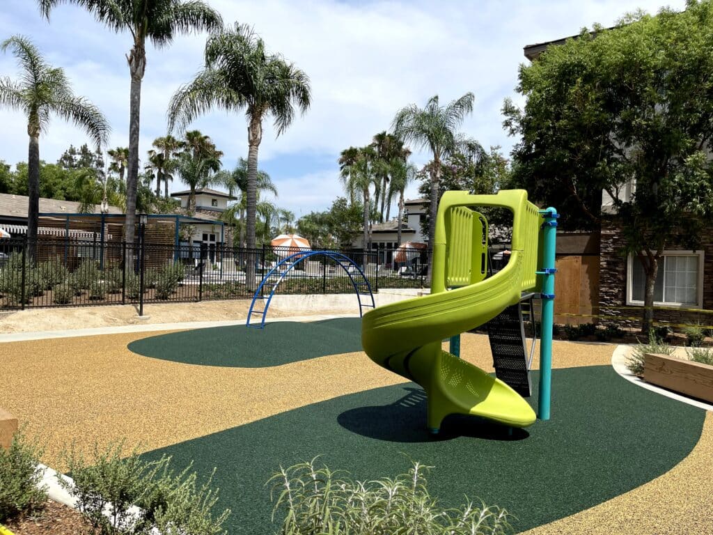 Dark green poured-in-place rubber play area with a lime-green spiral slide under palm trees