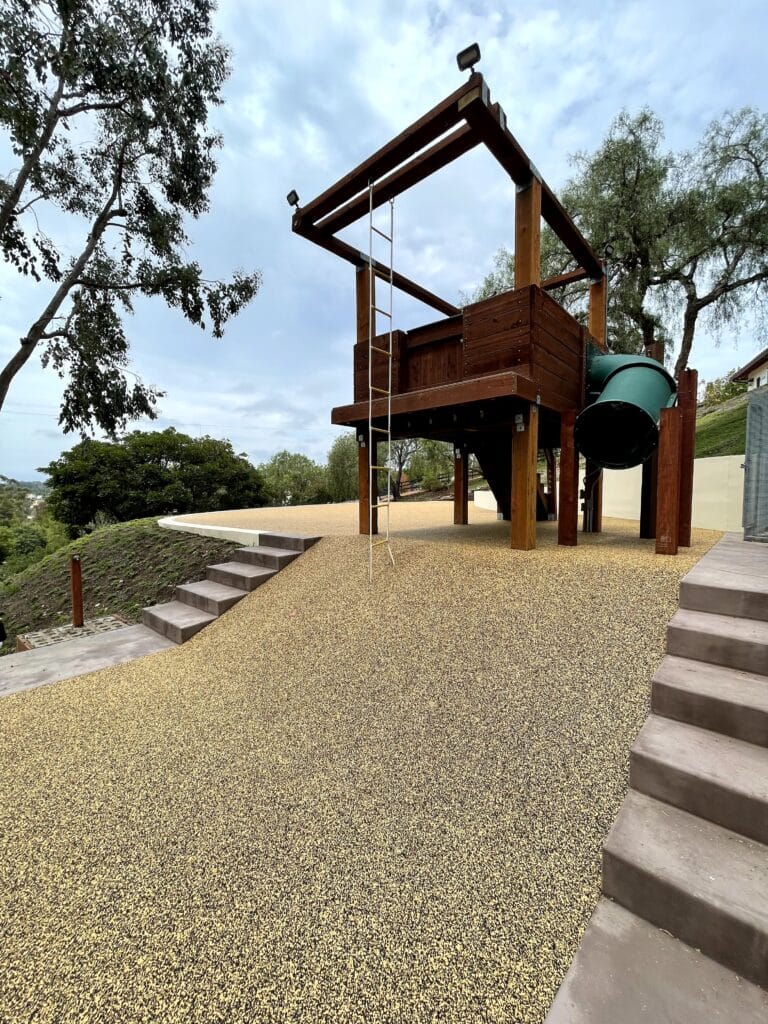 Porous rubber surfacing on a slope beneath a wooden play structure beside concrete steps