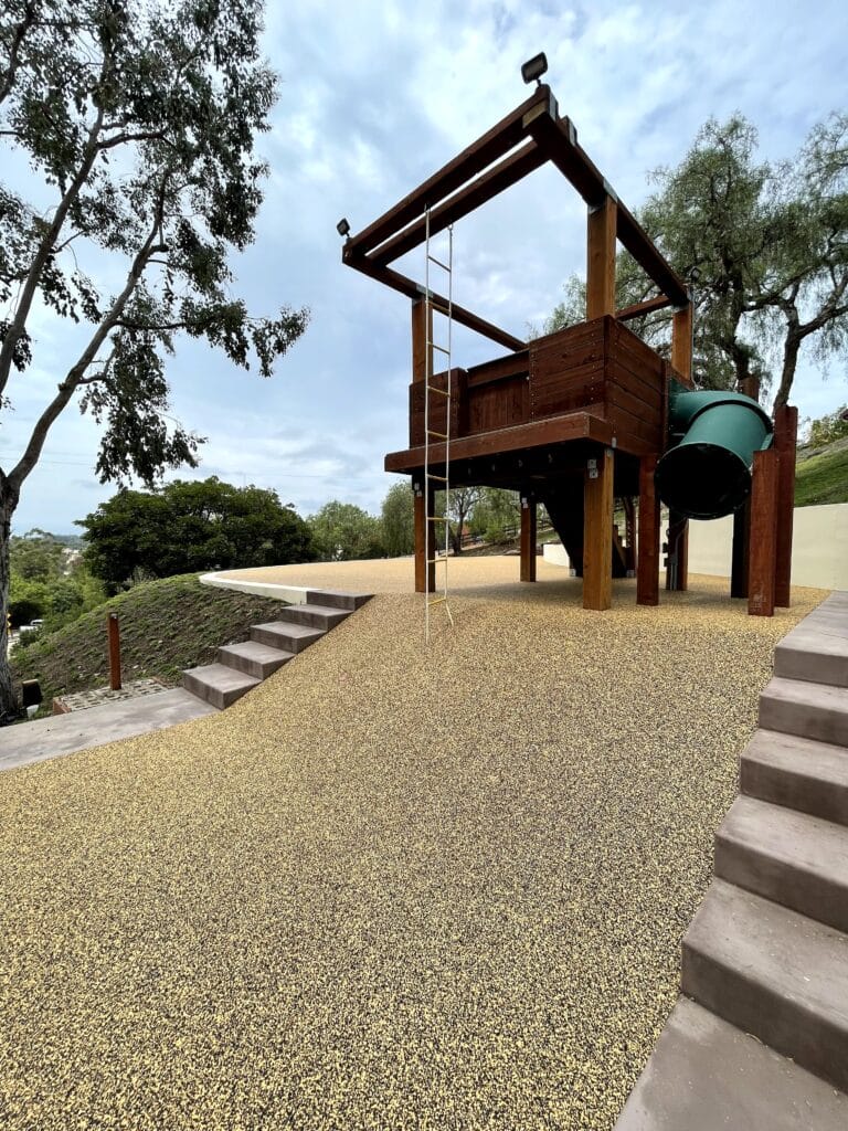 Tan porous rubber surface under a wooden play structure on a hillside