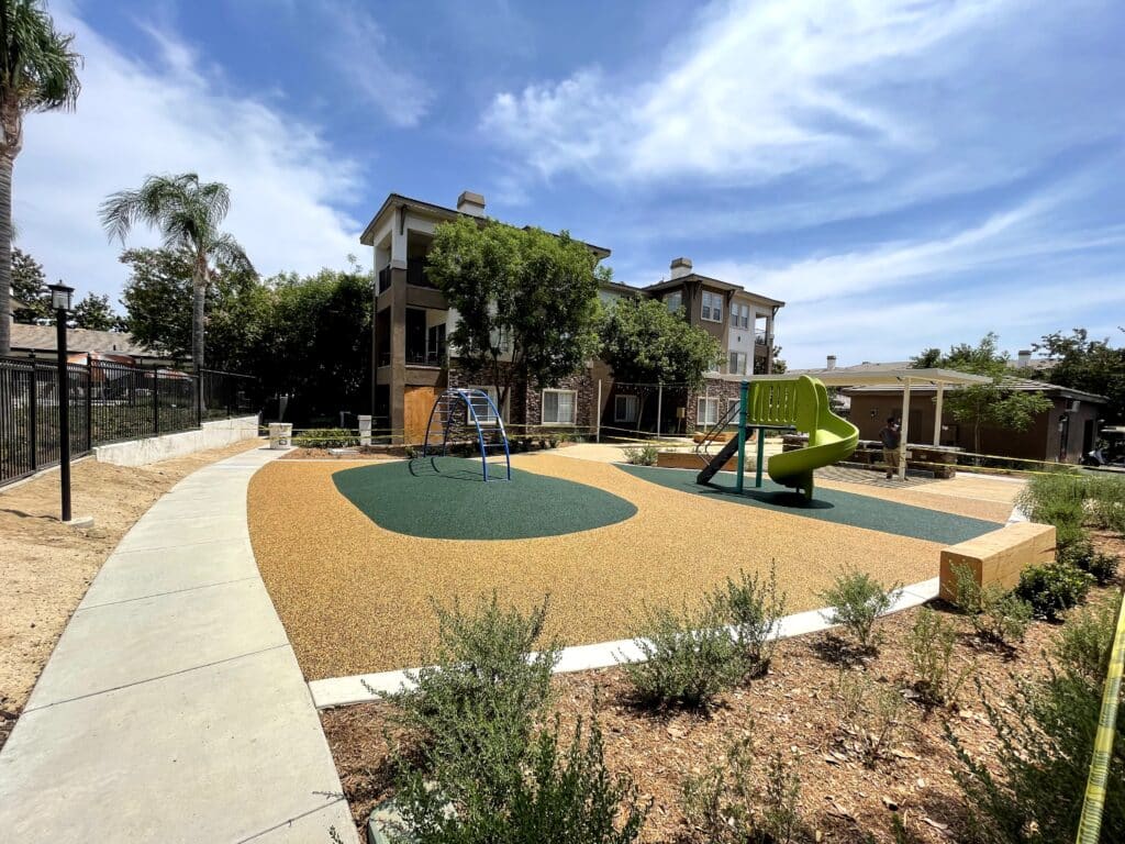Tan porous rubber surfacing around green play pads and play structures beside a walkway