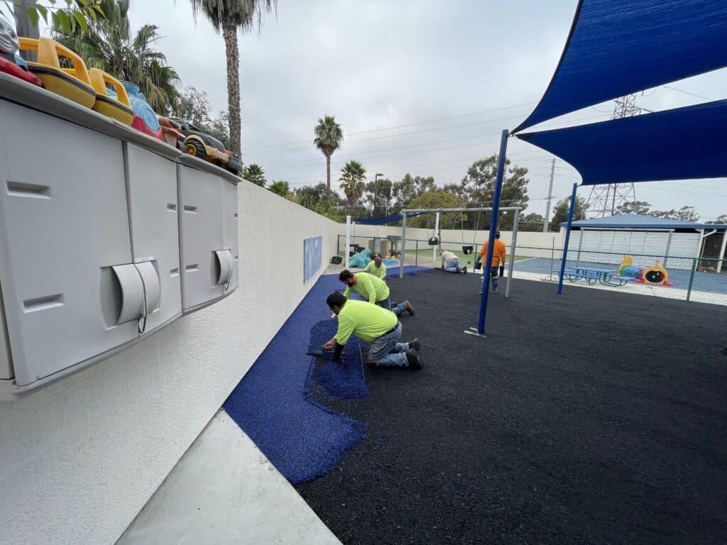 Installers placing poured-in-place rubber under a shade sail beside a building