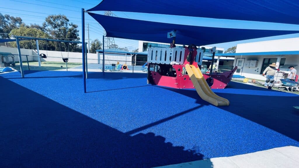 Blue and pink poured-in-place rubber under shade sails with a play structure and slide