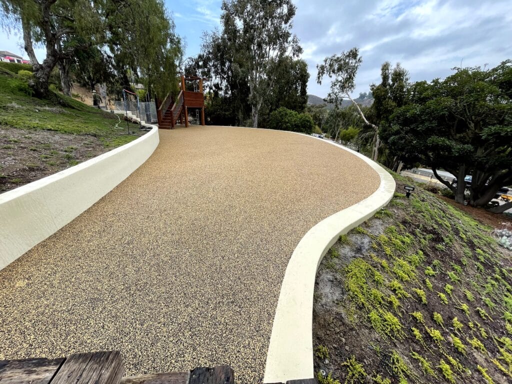Curving porous rubber pathway with a concrete curb on a hillside, with a play structure in the distance